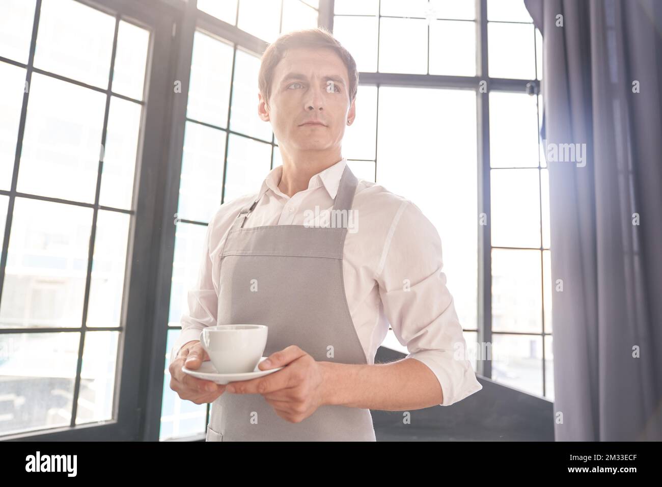 Ein männlicher Kellner in einer grauen Schürze hält eine Tasse Kaffee. Der Barista serviert eine Tasse heißen Kaffee vor dem Hintergrund eines großen Fensters mit Platz zum Kopieren. Hochwertiges Foto Stockfoto