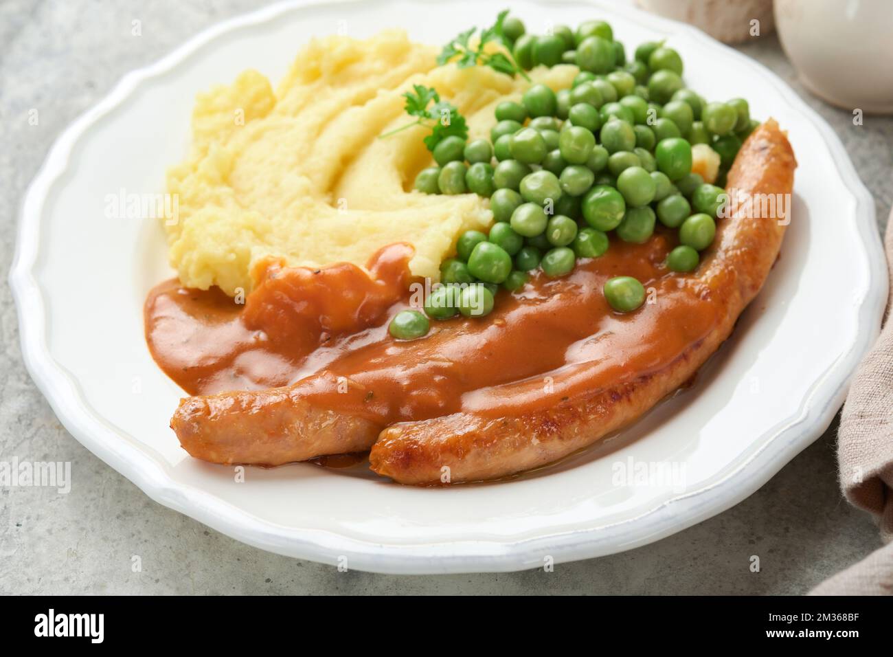 Knaller und Püree. Gegrillte Würstchen mit Kartoffelpüree und grüner Erbse auf weißem Teller auf grauem Hintergrund. Traditionelles Gericht aus Großbritannien und Irland. BB Stockfoto