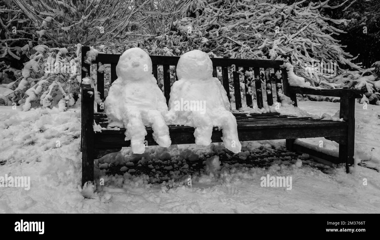 Schwarzweißbild von zwei Schneemännern, die nach dem ersten Schnee von 2022 auf einer Parkbank in London sitzen. Stockfoto