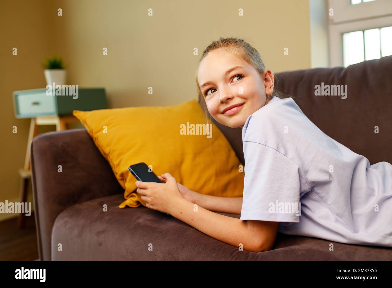 Süßes kleines Mädchen, das Smartphone benutzt und sich auf dem Sofa in ihrem viving-Zimmer zu Hause hinlegt Stockfoto