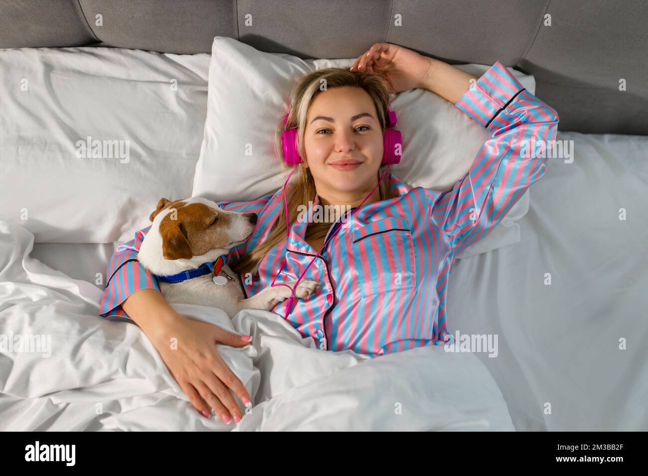 Eine sorgenfreie Frau im Pyjama, die Musik über Kopfhörer hört, während sie mit ihrem geliebten Haustier im Bett sitzt Stockfoto
