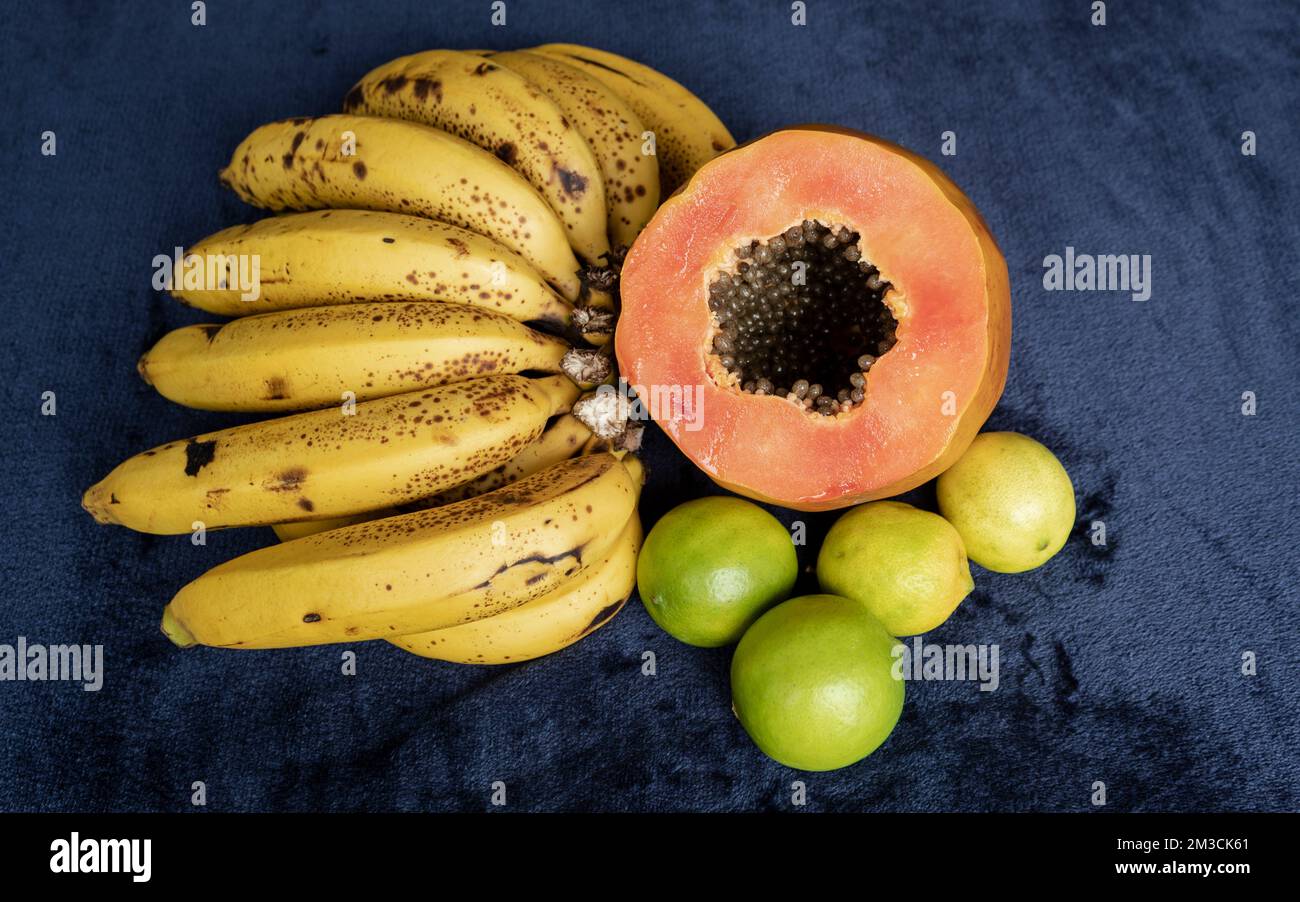 Draufsicht auf eine frisch geschnittene Orangenpapaya mit Samen im Inneren, eine Sommersprosse Bananen und drei grüne Zitronen auf blauem marineblauem und schwarzem Hintergrund. F Stockfoto
