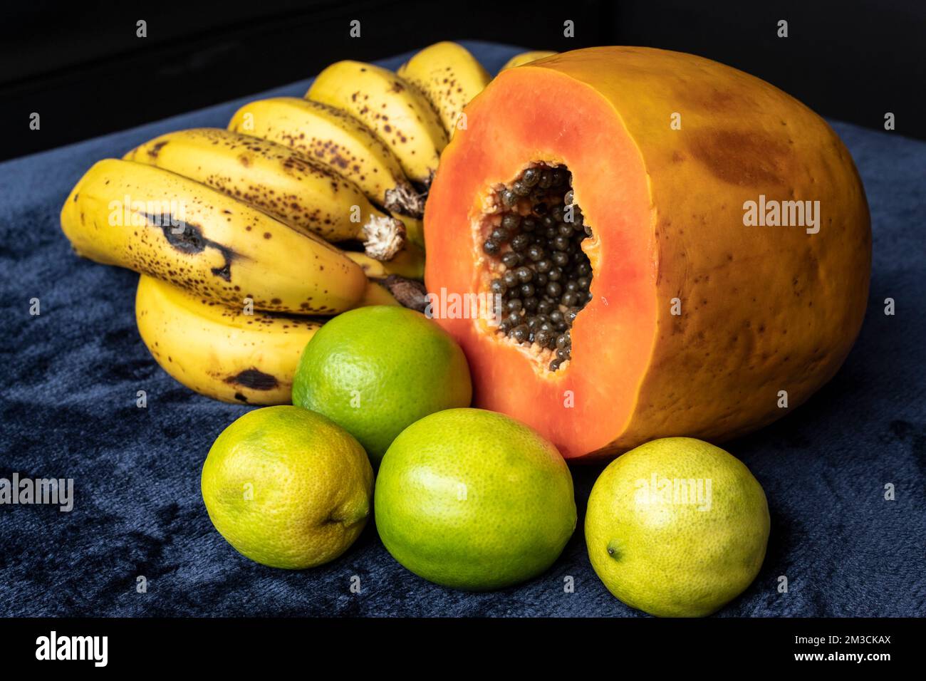 Aus nächster Nähe sehen Sie eine frisch geschnittene Orangenpapaya mit Samen im Inneren, eine Sommersprosse Bananen und drei grüne Zitronen auf blauem marineblauem und schwarzem Hintergrund. F Stockfoto