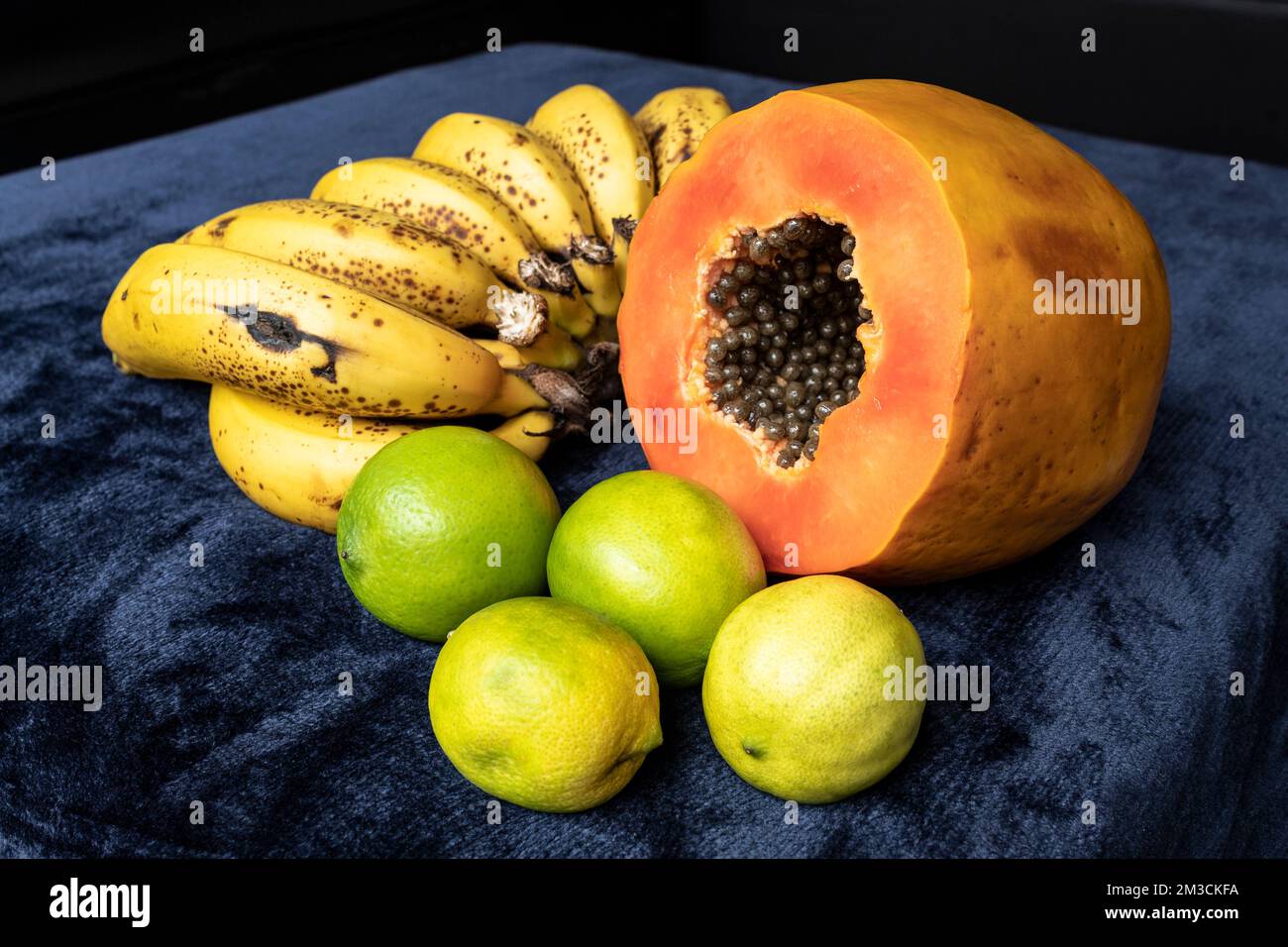 Aus nächster Nähe sehen Sie eine frisch geschnittene Orangenpapaya mit Samen im Inneren, eine Sommersprosse Bananen und drei grüne Zitronen auf blauem marineblauem und schwarzem Hintergrund. F Stockfoto