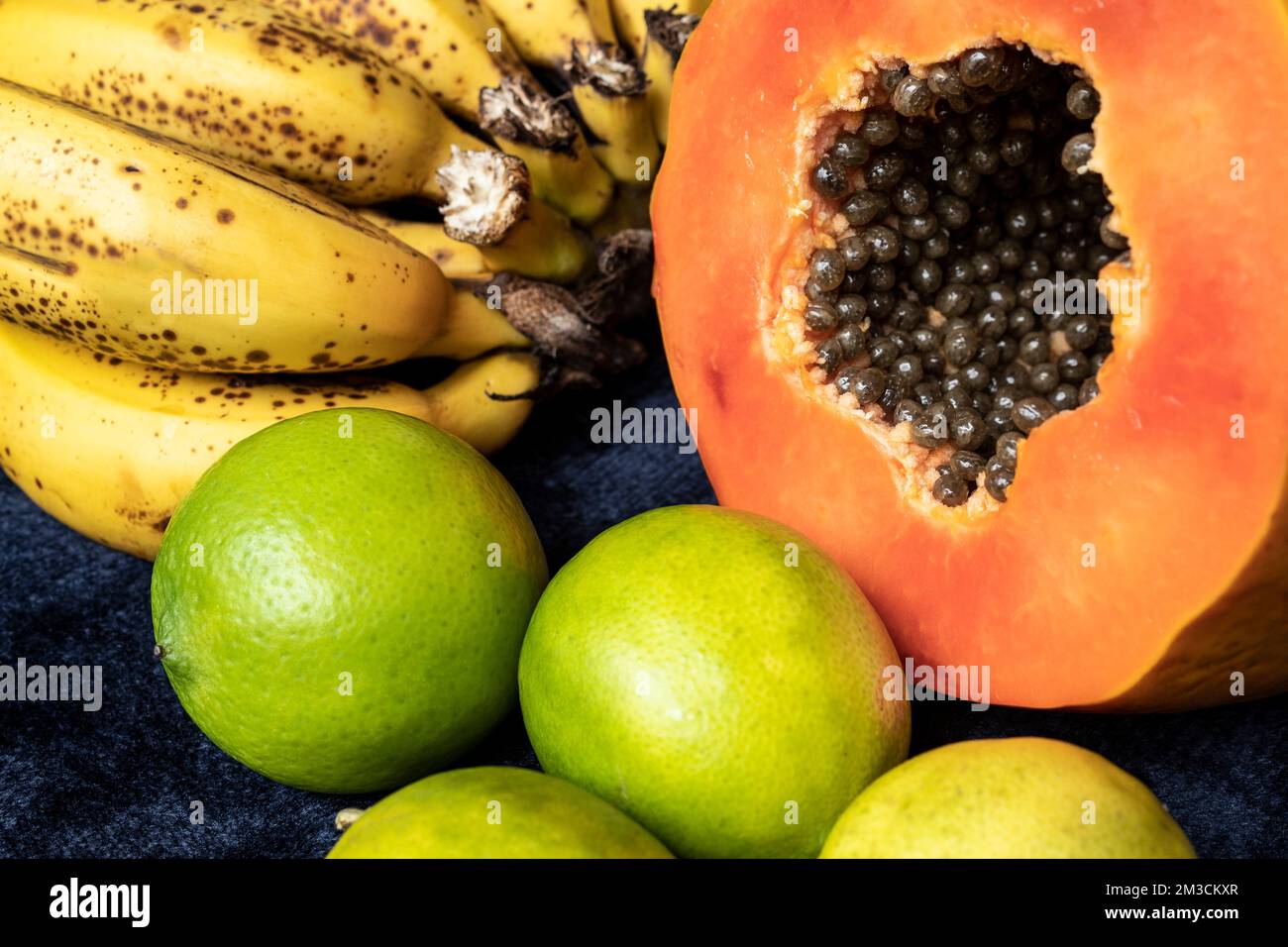 Aus nächster Nähe sehen Sie eine frisch geschnittene Orangenpapaya mit Samen im Inneren, eine Sommersprossen-Bananengruppe und drei grüne Zitronen auf blauem marineblauem Hintergrund. Obst und E Stockfoto