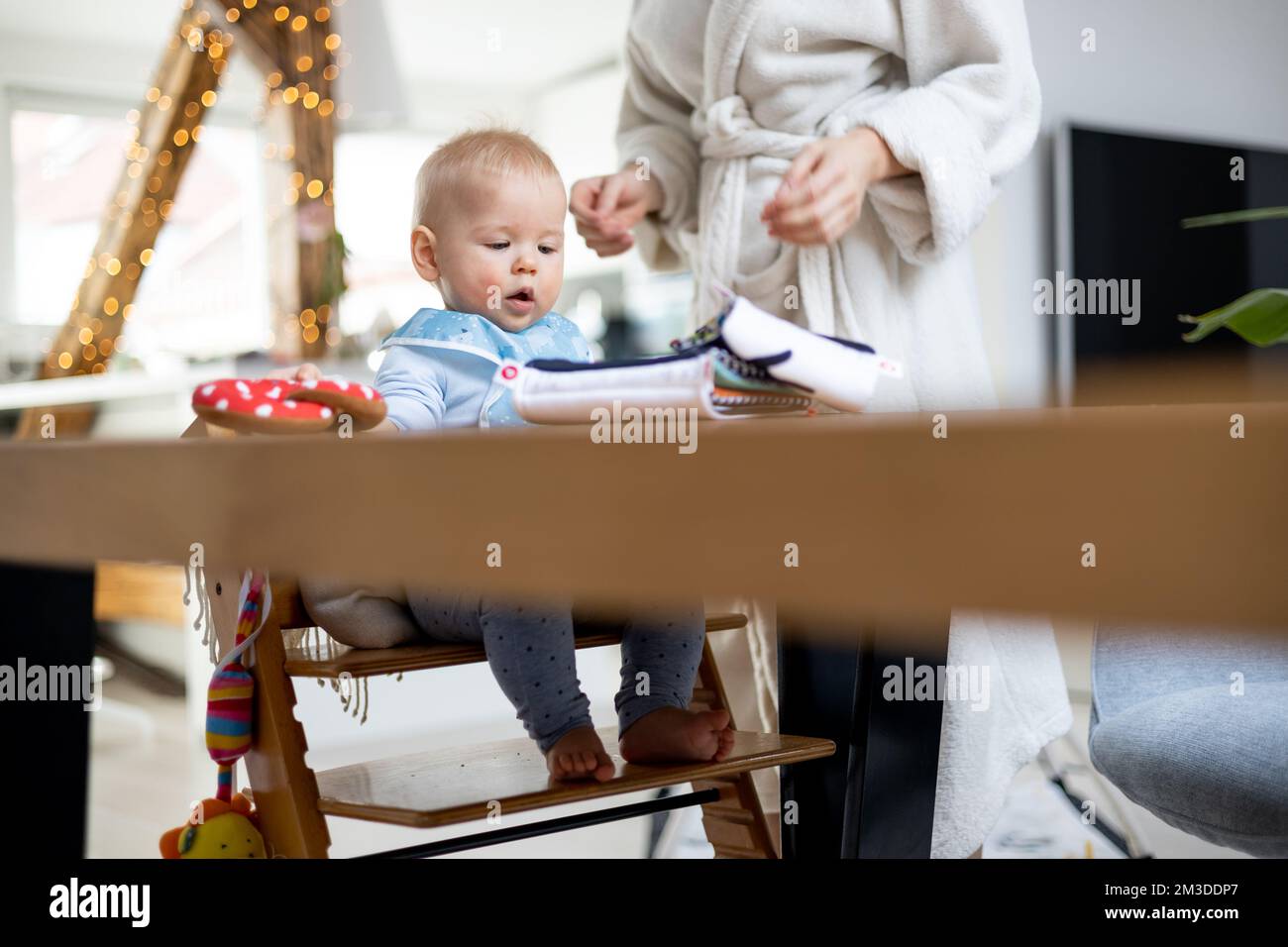 Glückliches Kind, das am Esstisch sitzt und mit seinem Spielzeug in einem traditionellen skandinavischen Designer-Hochstuhl aus Holz in einem modernen, hellen Haus spielt, das von seiner Mutter überholt wird. Stockfoto