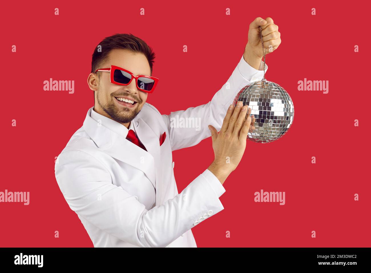 Ein fröhlicher, cooler junger Mann dreht eine glänzende kleine Discokugel isoliert auf rotem Hintergrund. Stockfoto