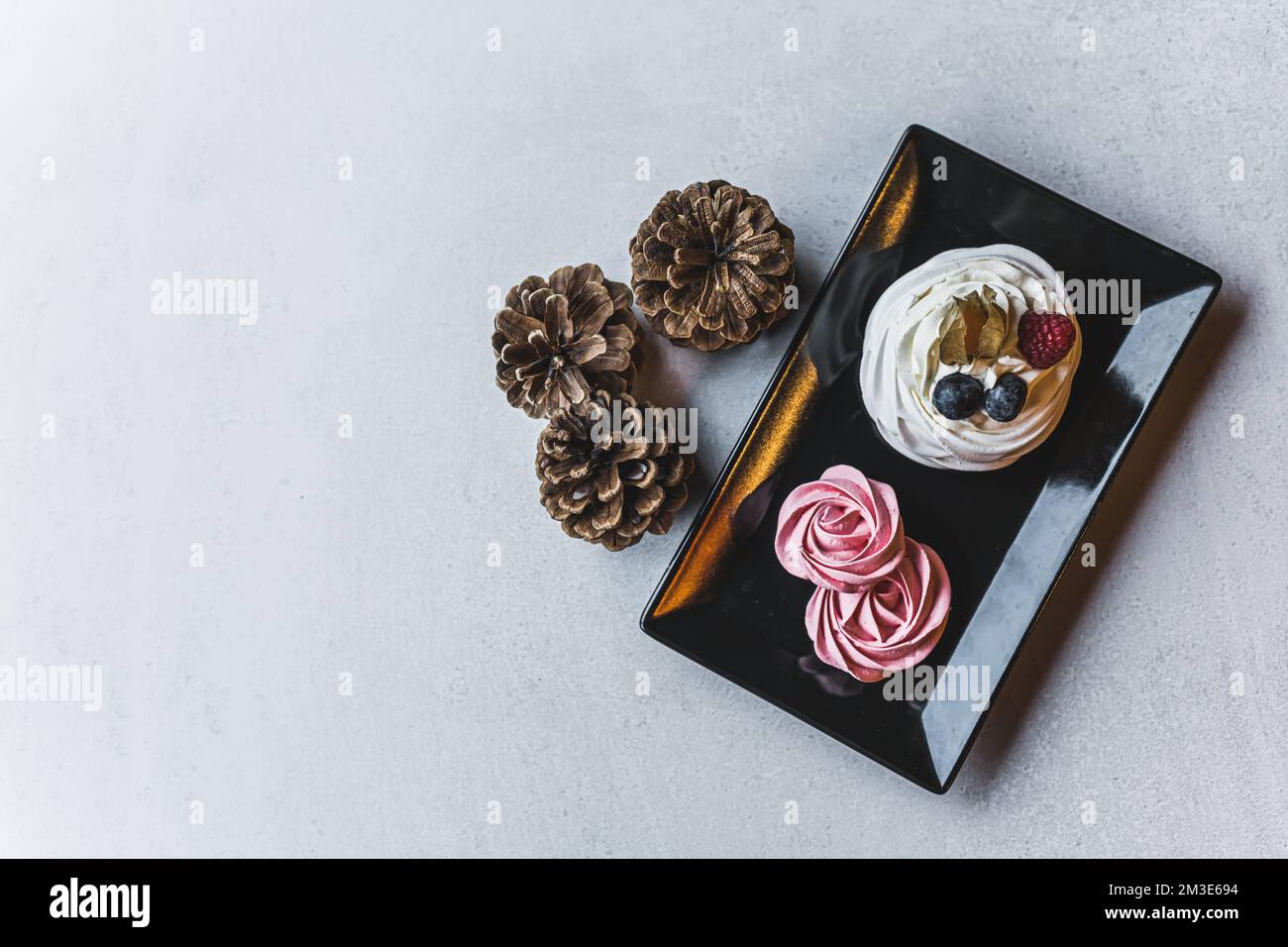 Schicke Baiser in Weiß und Pink auf einer schwarzen Platte auf einem weißen Tisch mit Pinienzapfen. Weihnachtsnachtisch. Professionelles Backen. Horizontale Draufsicht. Hochwertiges Foto Stockfoto