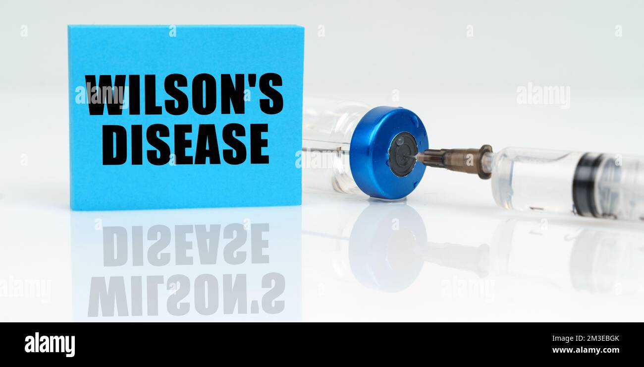 Medizinisches Konzept. Auf einer weißen reflektierenden Oberfläche sind eine Spritze, eine Injektion und eine blaue Plakette, auf der steht - Wilsons-Krankheit Stockfoto