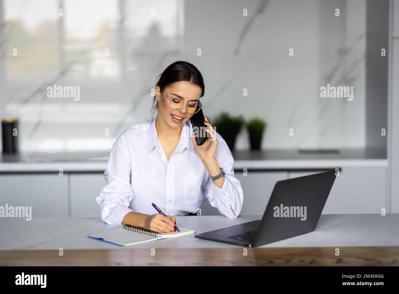 Aufnahme einer lächelnden Geschäftsfrau, die mit einem Notebook arbeitet, während sie zu Hause in der Küche mit einem Smartphone spricht. Stockfoto
