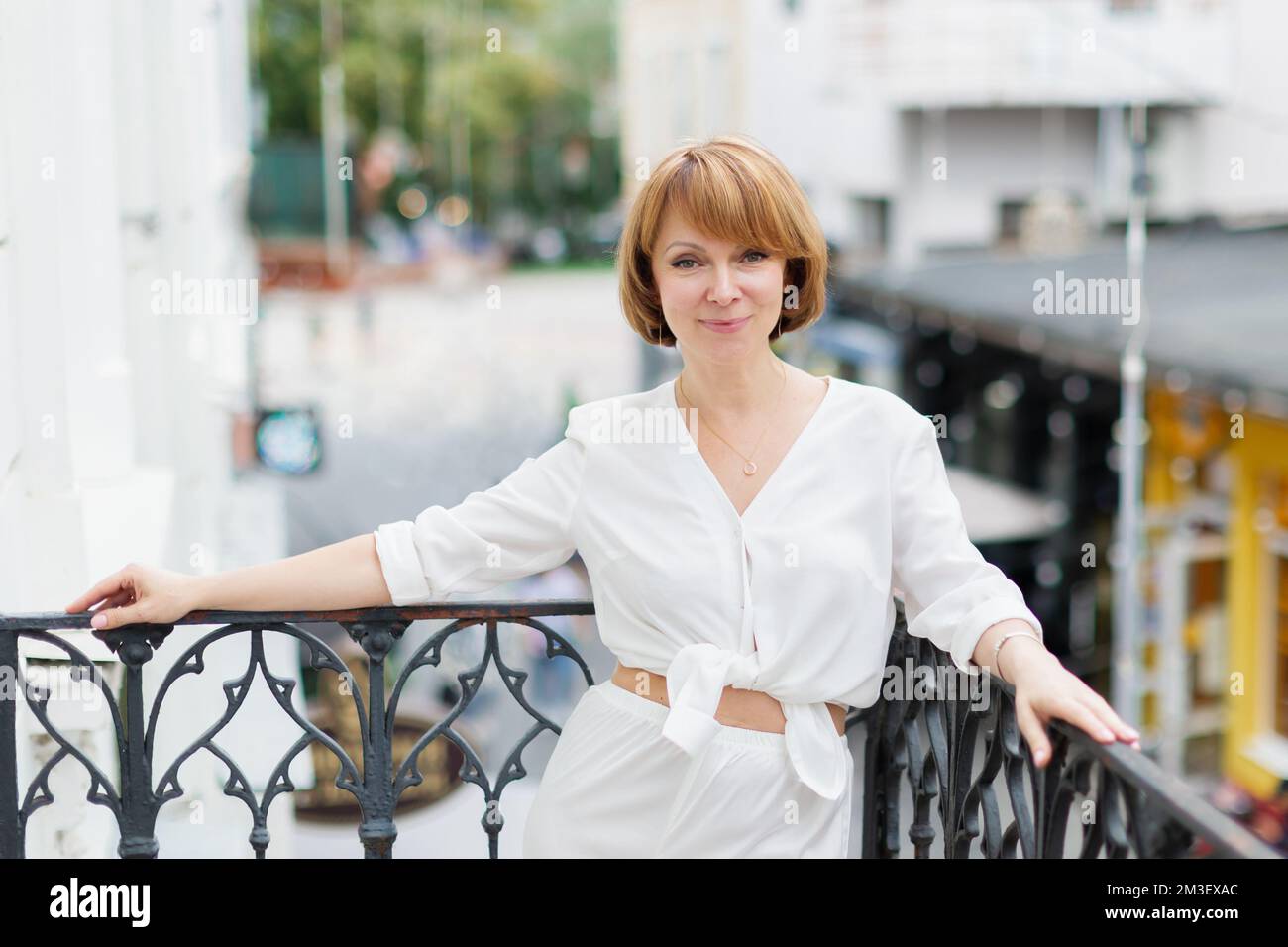 Erwachsene Frau in weißem Anzug steht auf dem Balkon in der Stadt und schaut auf die Kamera Stockfoto