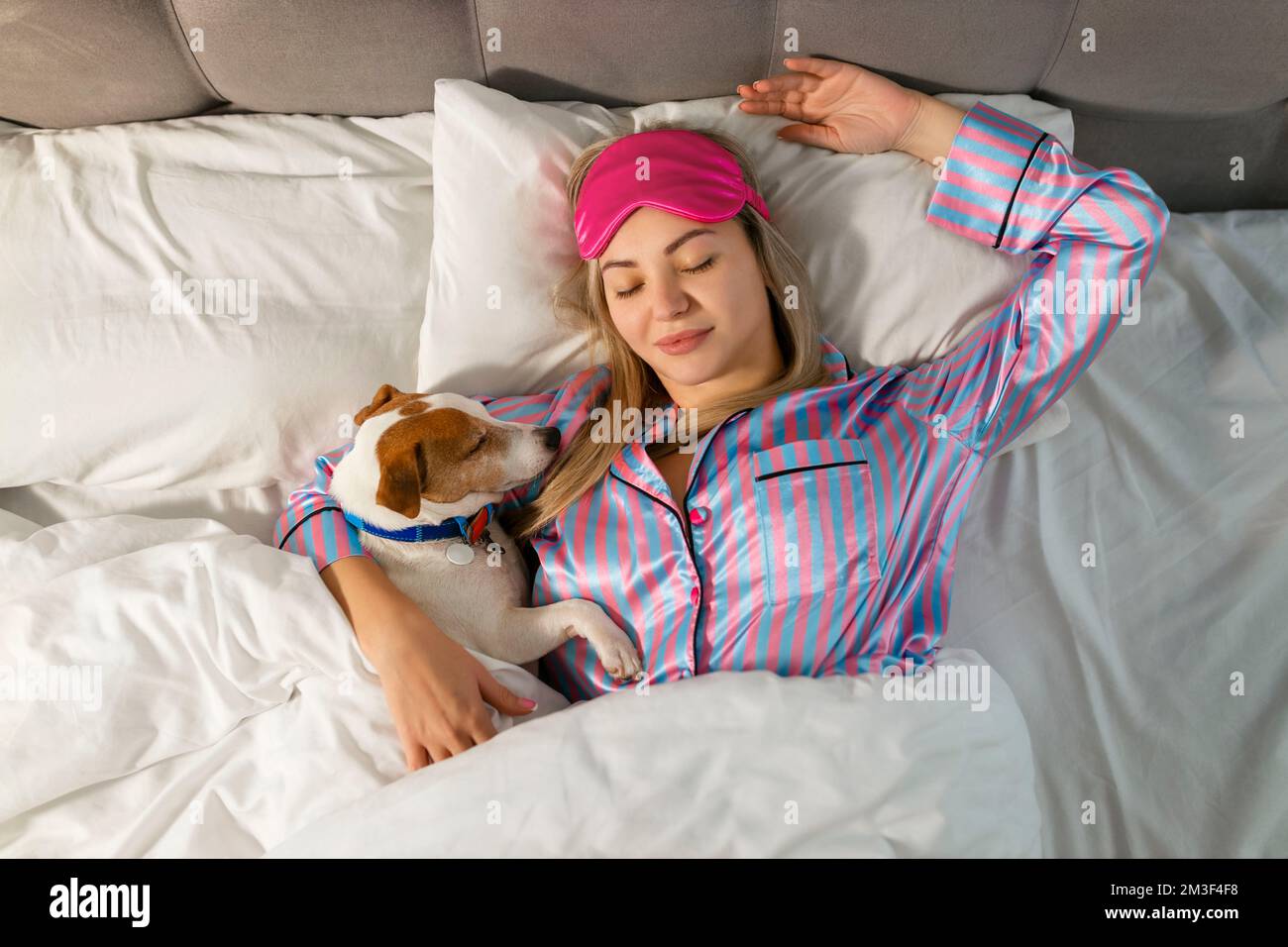 Eine attraktive junge Frau im Pyjama hält einen Hund, während sie auf einem Bett liegt Stockfoto