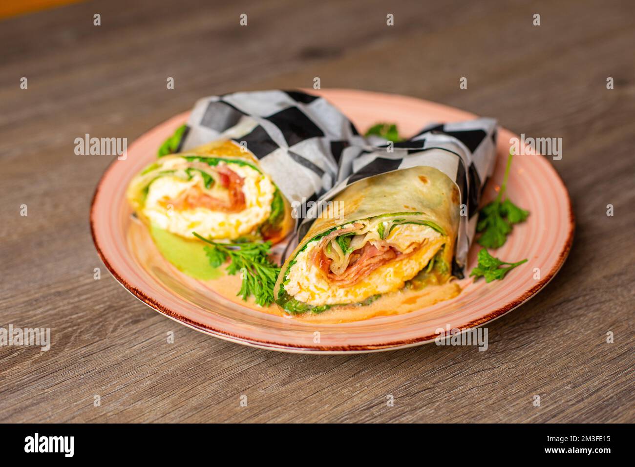 Leckere Speckbrötchen mit Grünzeug, Sauce und Schinken, verpackt in karierte Serviette, serviert auf einem Holztisch Stockfoto