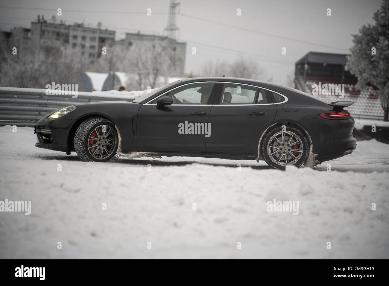 12-12-2022 Riga, Lettland ein schwarzer porsche parkt im Schnee vor einem Gebäude mit einem Uhrenturm im Hintergrund und einem Zaun im Vordergrund. Stockfoto