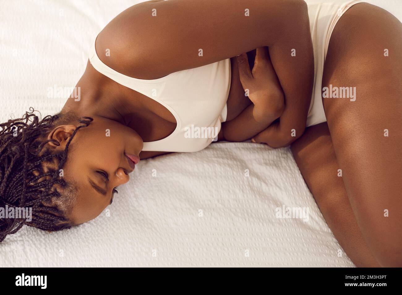 Junge Afroamerikanerin liegt auf dem Bett, hat Periodenkrämpfe und hält Hände auf dem Bauch Stockfoto
