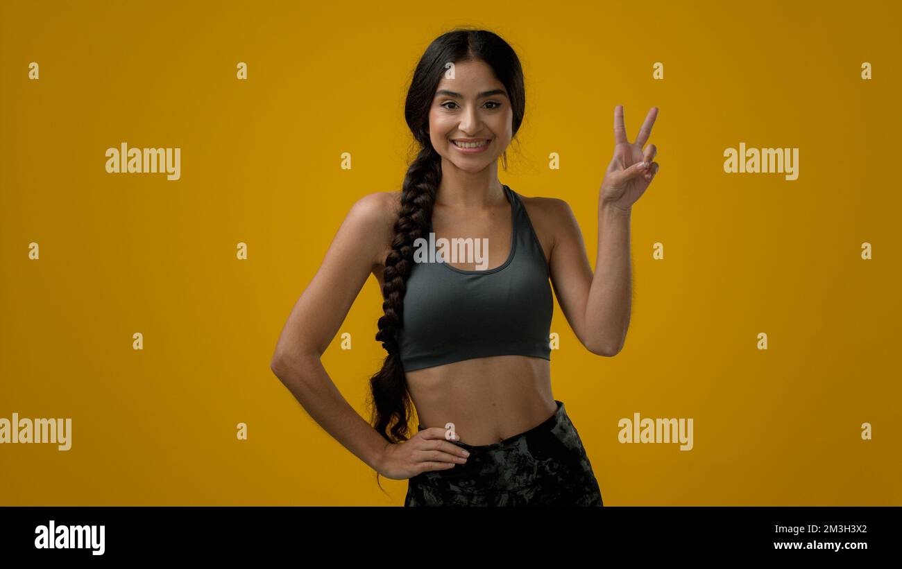 Lächelnde indische Sportlerin weibliche Sportlerin im gelben Studiohintergrund zeigt Doppelfinger Geste zwei 2-Nummer Sieg Zeichen Frieden Stockfoto