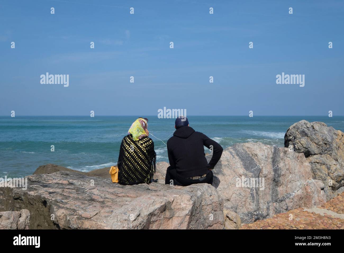 Muslimisches Paar, das auf dem Felsen sitzt, hört Musik und schaut auf das Meer, Rabat, Marokko. Stockfoto