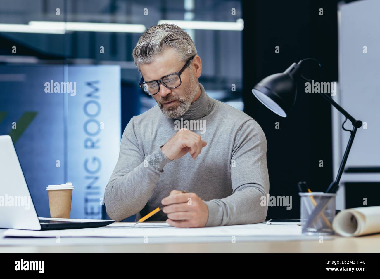 Konzentrierter männlicher Architekt, der abends mit einer Lampe im Büro arbeitet. Am Tisch sitzen, einen Plan mit einem Bleistift zeichnen, mit Papieren arbeiten. Stockfoto