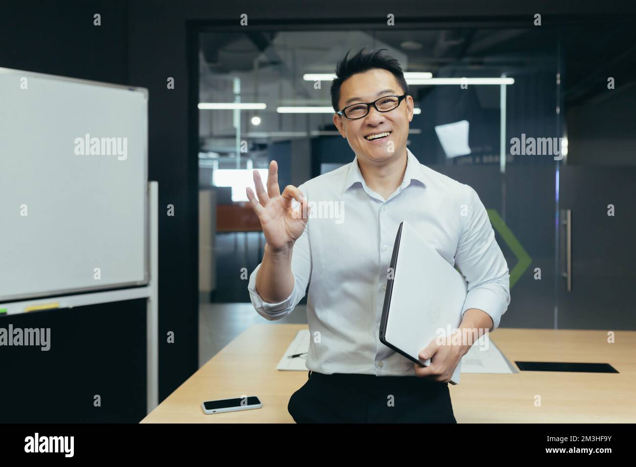 Porträt eines jungen, gutaussehenden asiatischen Studenten, Praktikanten, Assistenten. Er steht mit einem Ordner im Büro, trägt eine Brille, lächelt, zeigt mit den Fingern Stockfoto