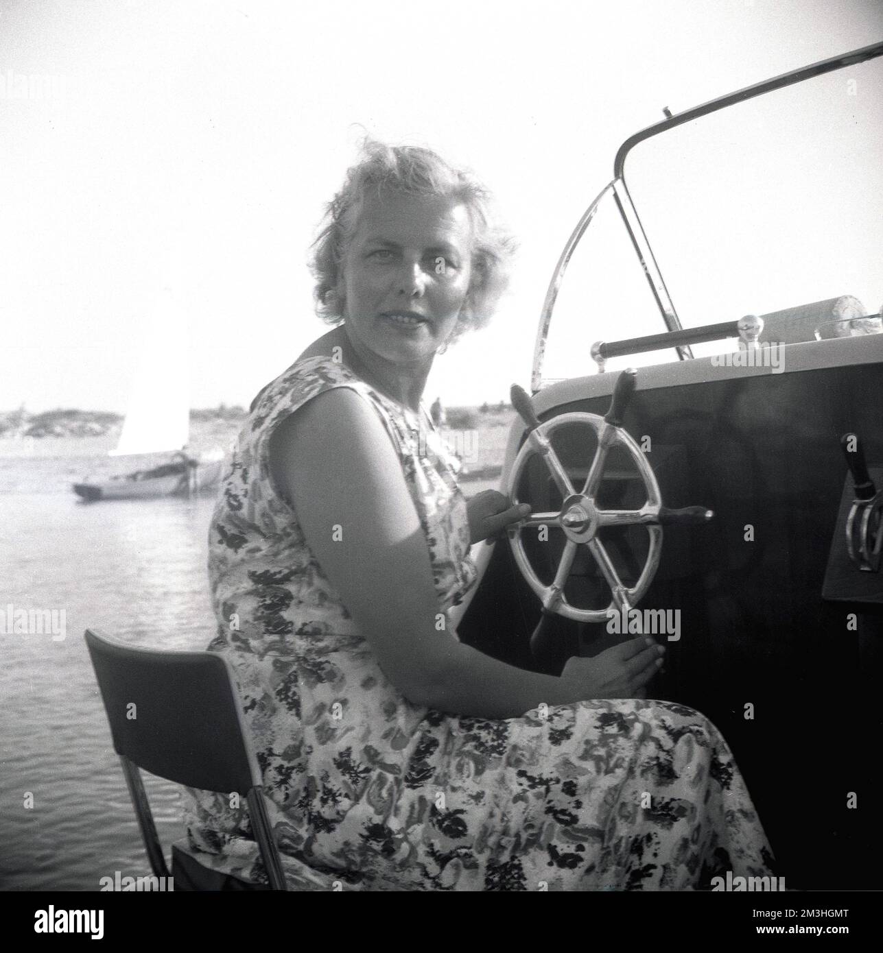 1960er, historisch, eine Dame sitzt am Steuer, T das Bootsrad eines Motorstarts, das in einem Hafen in England, Großbritannien, festgemacht ist. Stockfoto