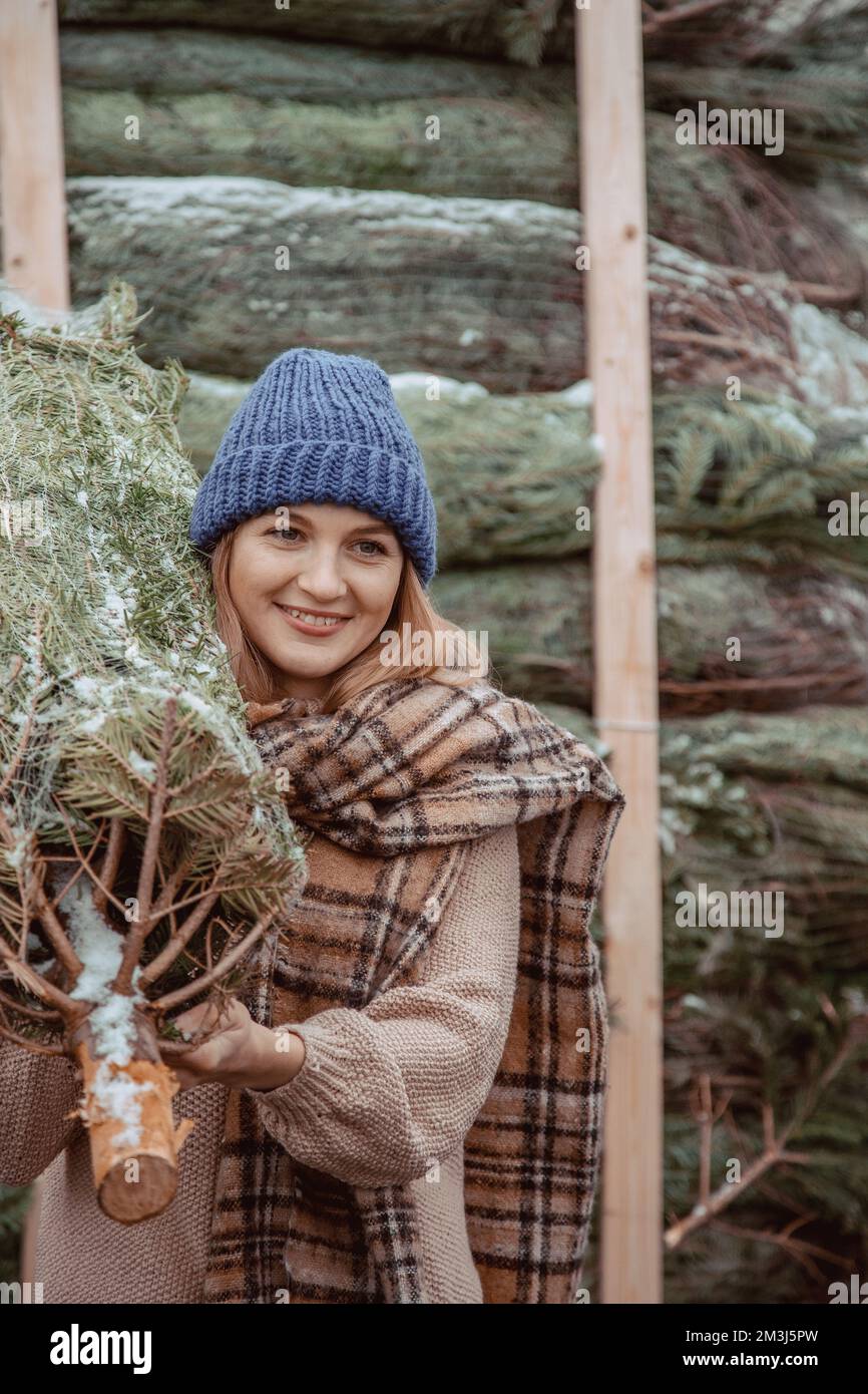 Ein hübsches Mädchen mit blauer Mütze und Schal trägt einen Weihnachtsbaum auf der Schulter. Ein Mädchen, das zu Weihnachten gekleidet ist, bringt eine frisch geschnittene Tanne mit nach Hause Stockfoto