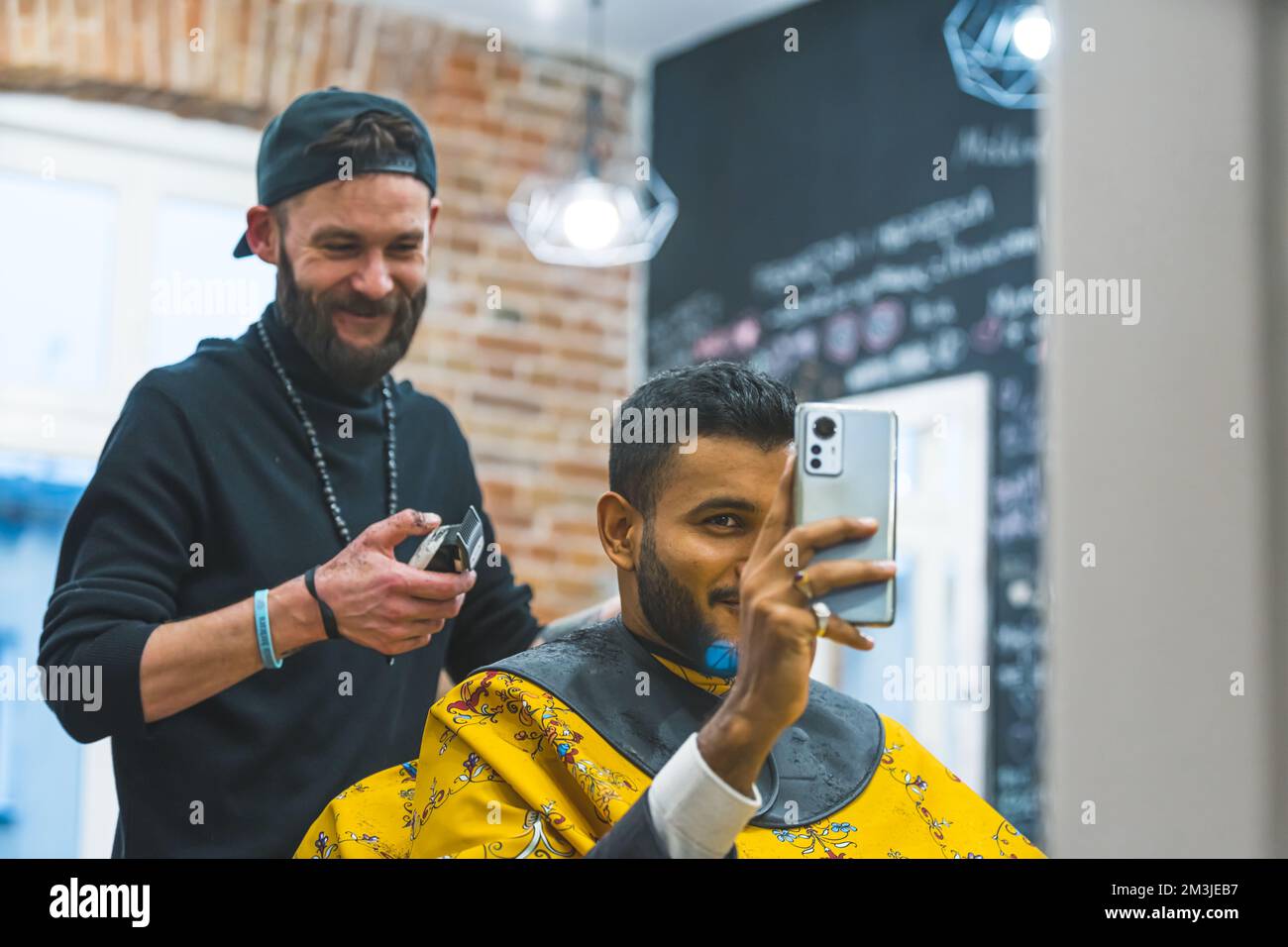 Ein moderner, eleganter Mann, der sich seine neue Frisur ansieht und sein Telefon als Spiegel benutzt. Ein gutaussehender indischer Influencer macht ein Selfie im Friseursalon. Hochwertiges Foto Stockfoto