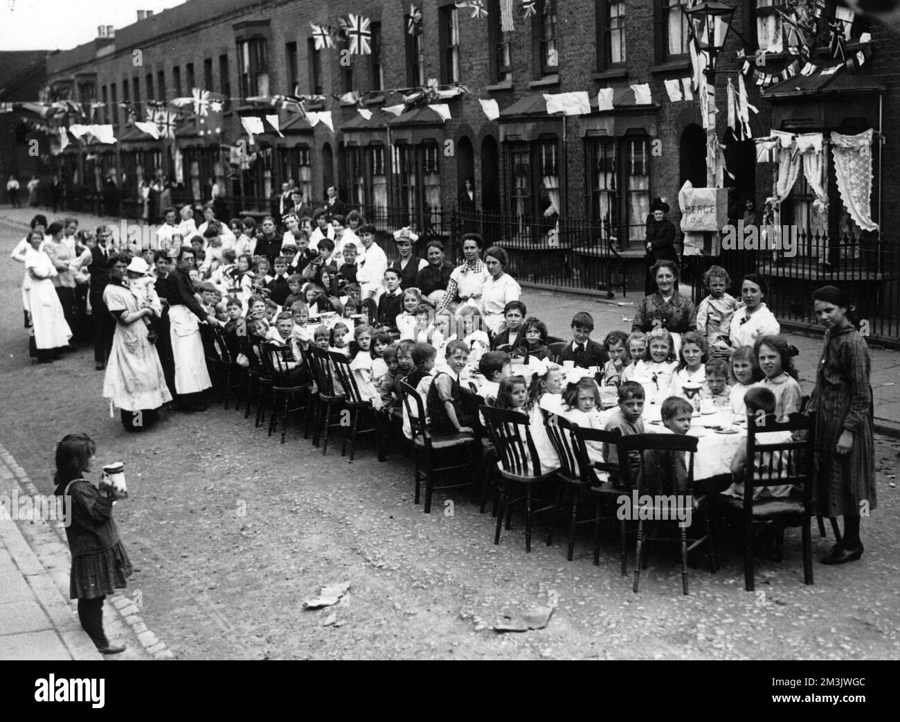 Teeparty in einer Straße in Londons East End, um das Ende des Ersten Weltkriegs im Jahr 1918 zu feiern. Kinder sitzen an einem langen Tisch, während Eltern (hauptsächlich Frauen) Essen servieren. Bunting, Flaggen und Banner sind entlang der Straße zu sehen. Datum: 1918 Stockfoto