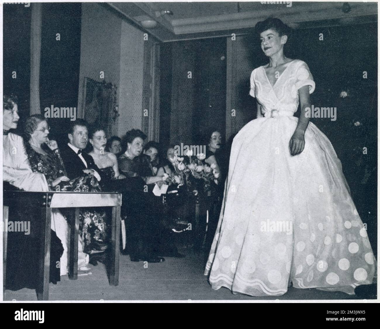 Ein weißes Abendkleid aus organischem Anbau mit großen weißen Punkten. Die schlanke Taille wurde durch die Fülle des Rocks betont. 1947 hat Christian Dior eine sensationelle Kollektion mit dem Titel „New Look“ entworfen. Seine Designs konzentrierten sich auf Kleider mit einer Taille von ca. 18 cm, die die Aufmerksamkeit auf Brust und Hüfte lenkten. Madame Maggy Besancon de Wagner, wie viele Designer, hat sich von Diors Sammlung inspirieren lassen. Ihre Modenschau im Frühling 1948 folgte der 18 cm langen Taille und dem Hüftdesign. Stockfoto