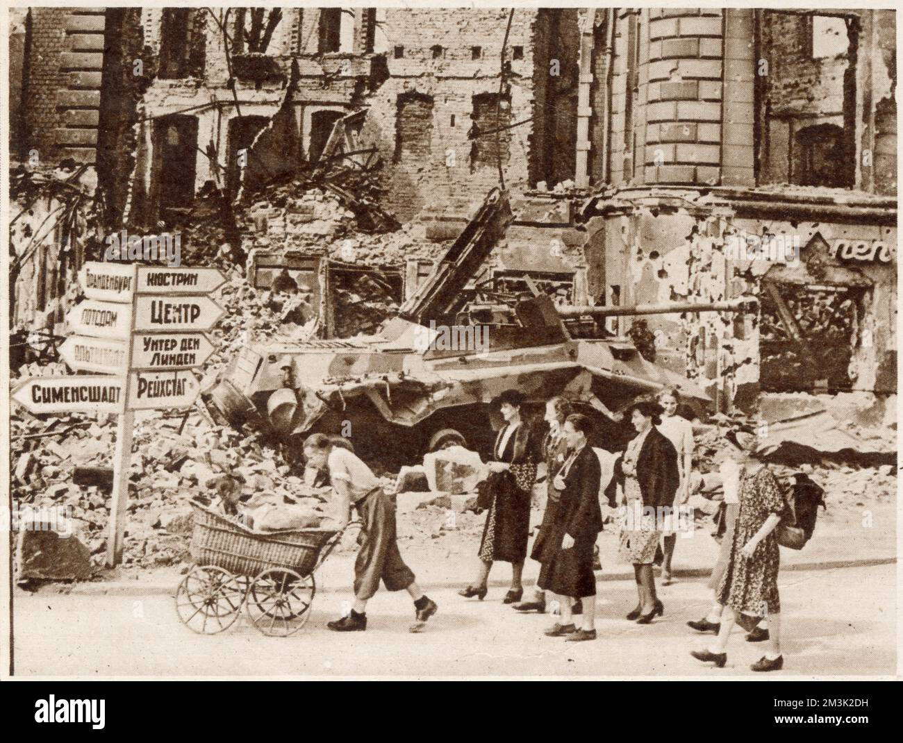 Eine Gruppe von Berlinern, die an den Ruinen des Nollendorf-Platzes vorbeispazieren und nach dem Ende des Zweiten Weltkriegs, Sommer 1945, nach einem Ort zum Leben suchen. Im Hintergrund stehen ein zerstörter deutscher Panzer und ein russisches Straßenschild nebeneinander. Stockfoto