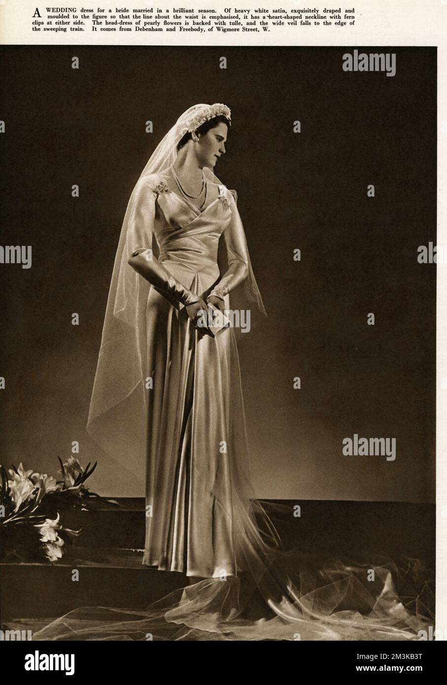 Schweres weißes Hochzeitskleid aus Satin, exquisit drapiert und an die Figur angepasst, sodass die Linie an der Taille betont wird. Es hat einen herzförmigen Ausschnitt mit Farnclip an beiden Seiten. Das Kopfkleid der Perlblüten ist mit Tüll bedeckt, und der breite Schleier fällt an den Rand des Zuges. 1937 Stockfoto