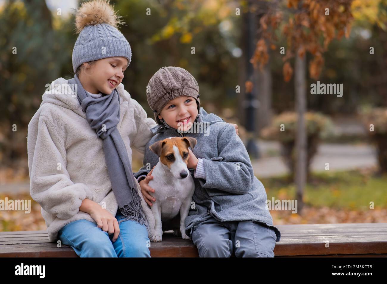 Bruder und Schwester sitzen in einer Umarmung mit einem Hund auf einer Bank für einen Spaziergang im Herbstpark. Junge, Mädchen und Jack russell Terrier. Stockfoto