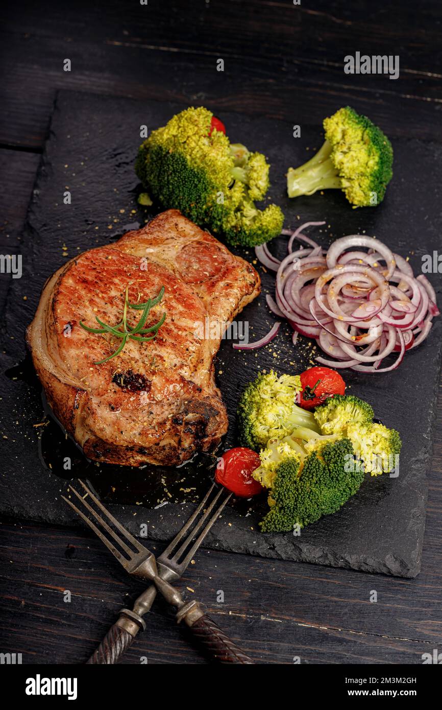 Schmackhaftes BBQ-Schweinelende mit Gemüse auf einem schwarzen Steinbrett. Schweinebraten und Brokkoli. Vertikaler Schuss. Speicherplatz kopieren. Altrustikaler Holzhintergrund Stockfoto