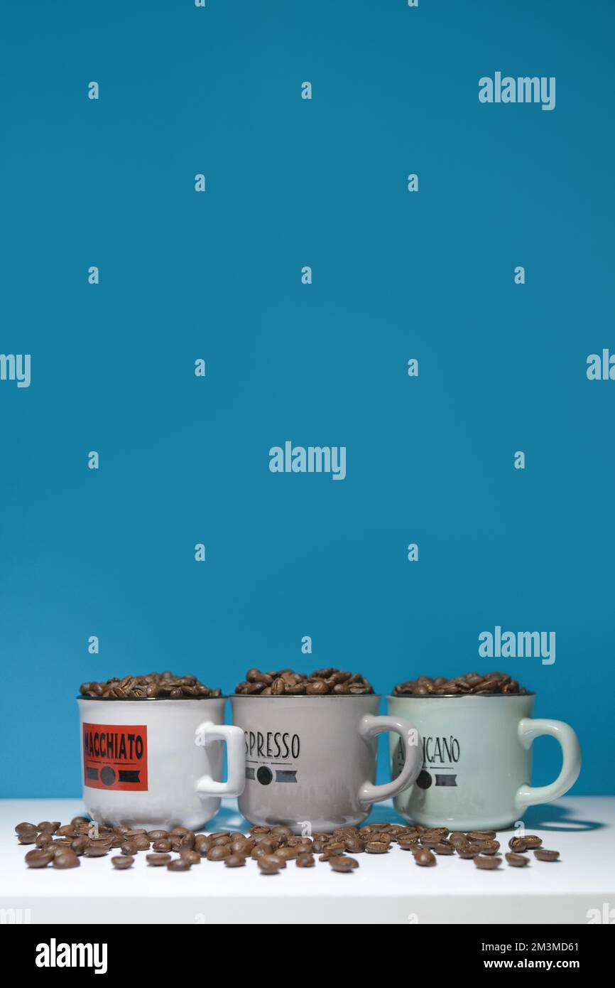 Espresso und mehr. Macchiato americano. Kaffeekörner in Tassen mit verschiedenen Namen auf blauem Hintergrund vertikal Stockfoto