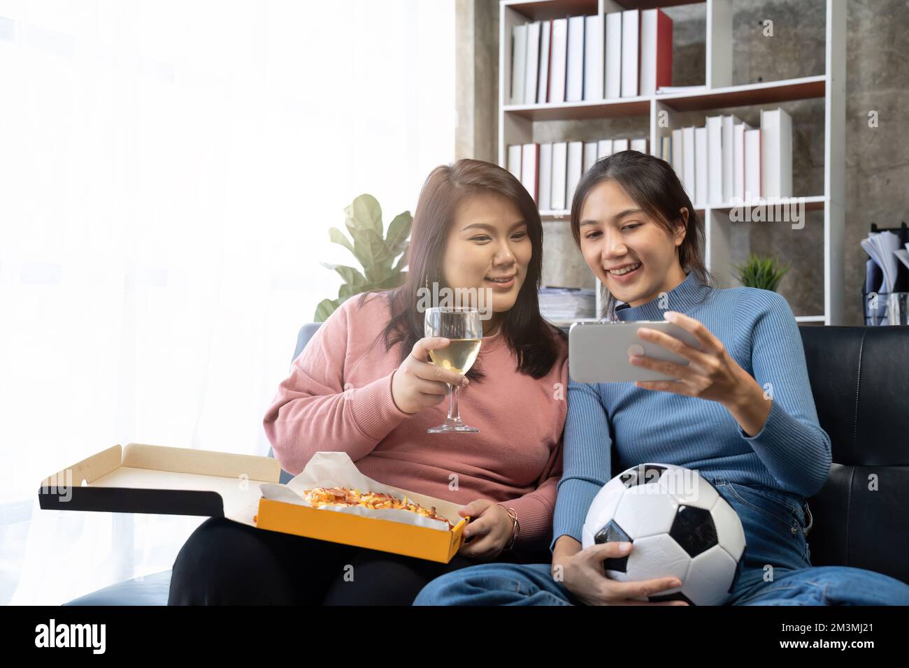 Freunde sehen sich Sport auf dem Handy an, jubeln und feiern. Fröhliche, vielfältige asiatische Fans setzen sich mit Popcorn und Drinks auf die Couch Stockfoto