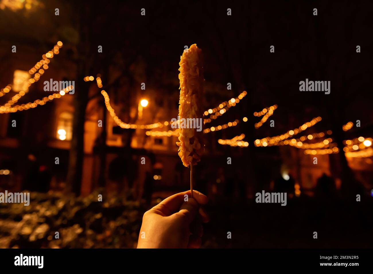 In einer Hand auf einem Holzstock ein Maishund in Flocken vor dem Hintergrund einer urbanen Weihnachtsdekoration. Verschwommenes orangefarbenes Bokeh. Straßenküche auf einem Fest Stockfoto