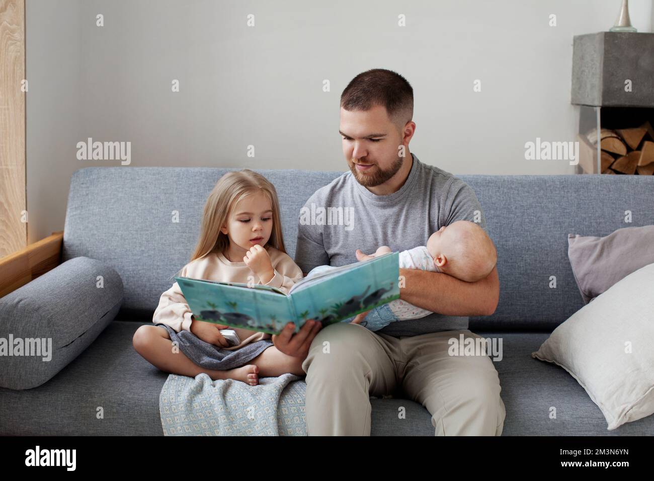 Junger alleinerziehender Vater mit zwei Kindern, die zu Hause auf der Couch sitzen, seiner Tochter ein Buch vorlesen und das Neugeborene in den Armen halten. Zeit für die Familie Stockfoto