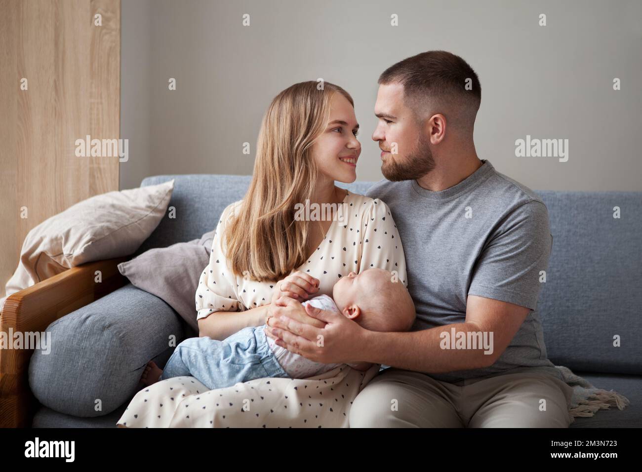 Glückliche Familie mit neugeborenem Baby. Eltern, eltern, eltern, eltern, halten ihr Baby in den Armen, lächeln und schauen sich an, wie sie zu Hause auf der Couch sitzen Stockfoto