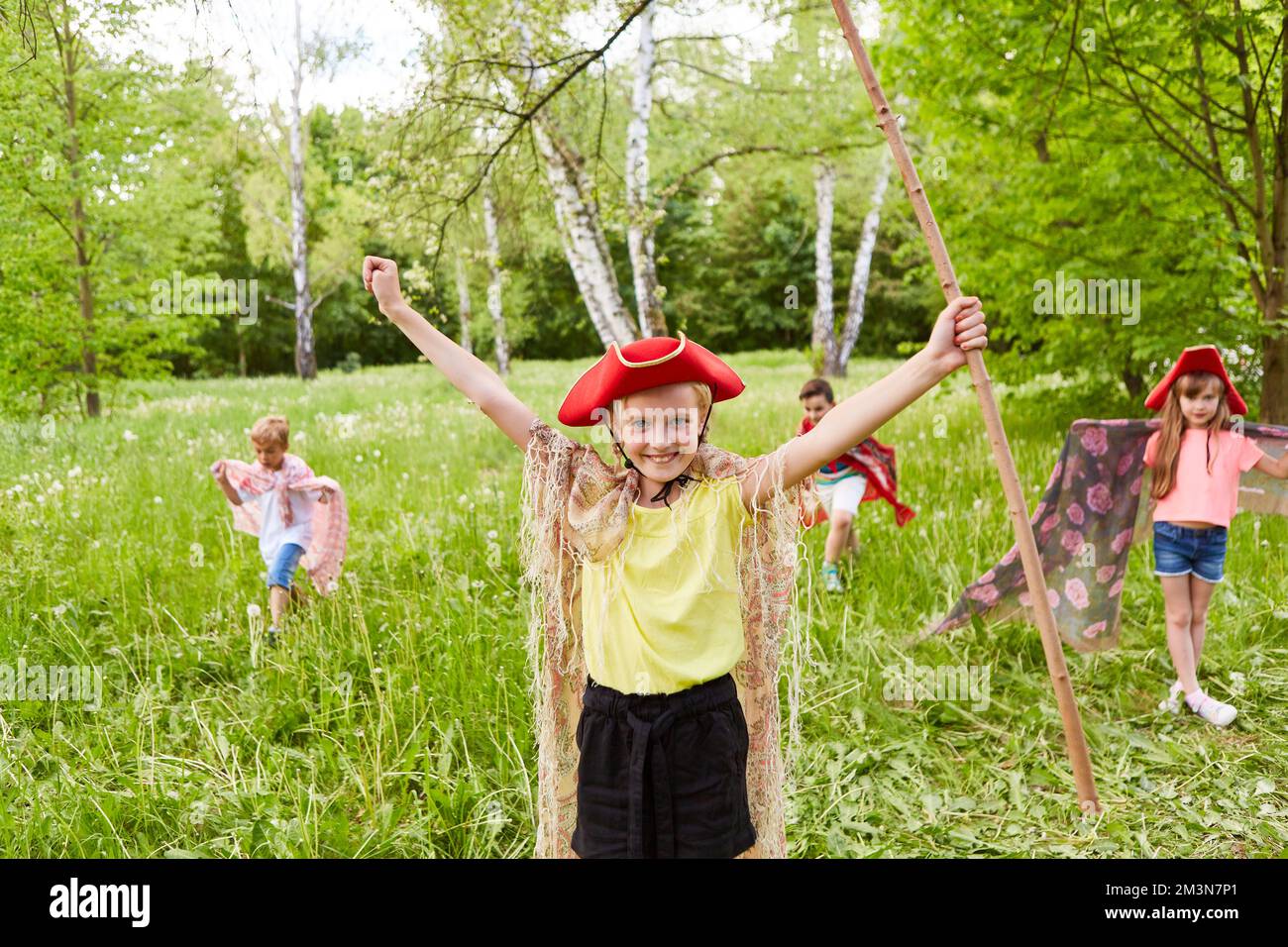Lächelndes Mädchen mit Tricornhut und Stock, die Arme von Freunden ausgestreckt im Gras Stockfoto