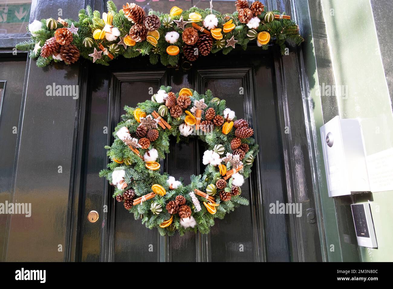 Wunderschöner Weihnachtskranz mit Kiefernzapfen, Zimtstiften und orangefarbenen Scheiben, die an der grünen Tür hängen, London England, UK, KATHY DEWITT Stockfoto
