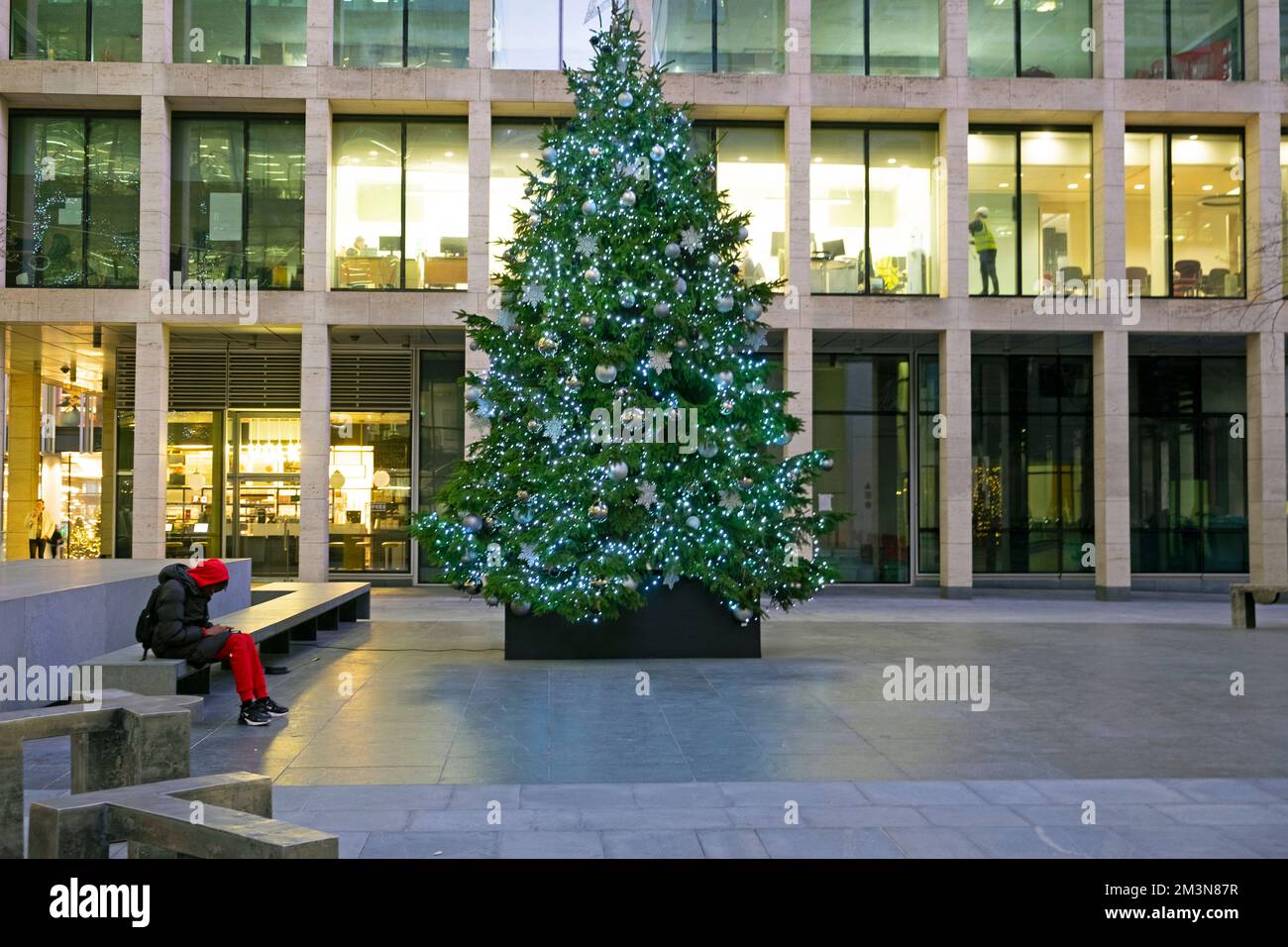 Person trägt rot, sitzt auf einer Bank am Bürogebäude des Weihnachtsbaums im Winter New Street Square London EC4 UK England Großbritannien KATHY DEWITT Stockfoto