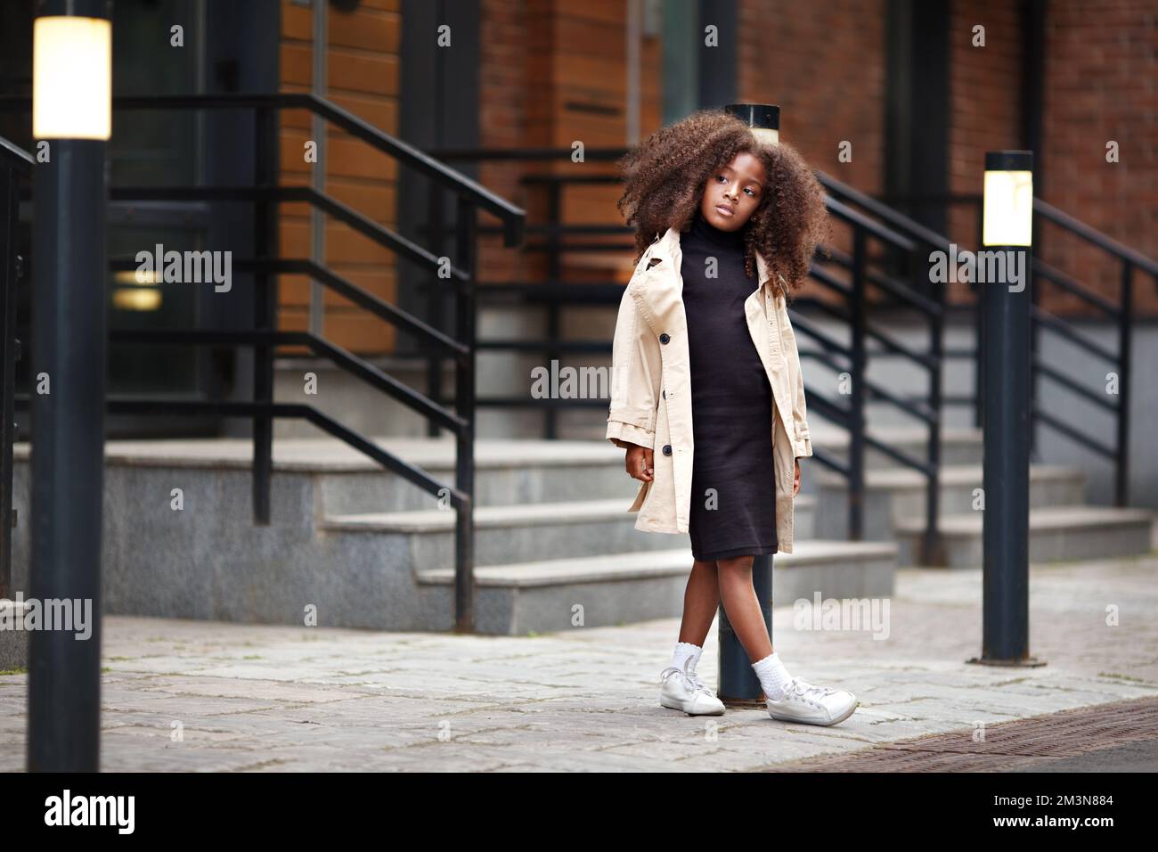 Ein nachdenkliches kleines Mädchen, das auf der Straße steht und auf jemanden wartet, wegschaut, modisch gekleidet in Trenchcoat und schwarzem Kleid. Volle Länge po Stockfoto