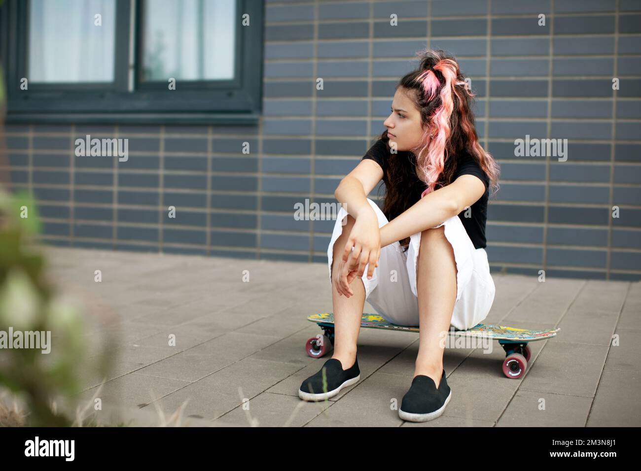 Teenager-Mädchen sitzt auf Skateboard. Hipster Teen Skateboarder mit langen pinkfarbenen Haaren auf grauem Hintergrund Stockfoto