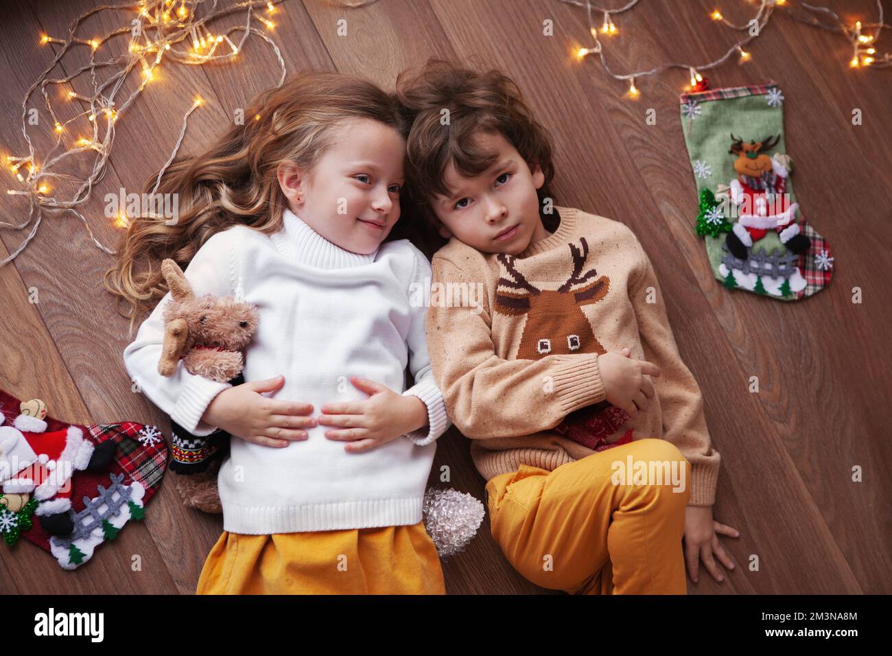 Porträt von süßen Kindern, die zu Hause auf dem Boden liegen, innen mit Weihnachtsdekor, Girlande aus Glühbirnen und Socken. Der schöne Junge und das Mädchen tragen S Stockfoto