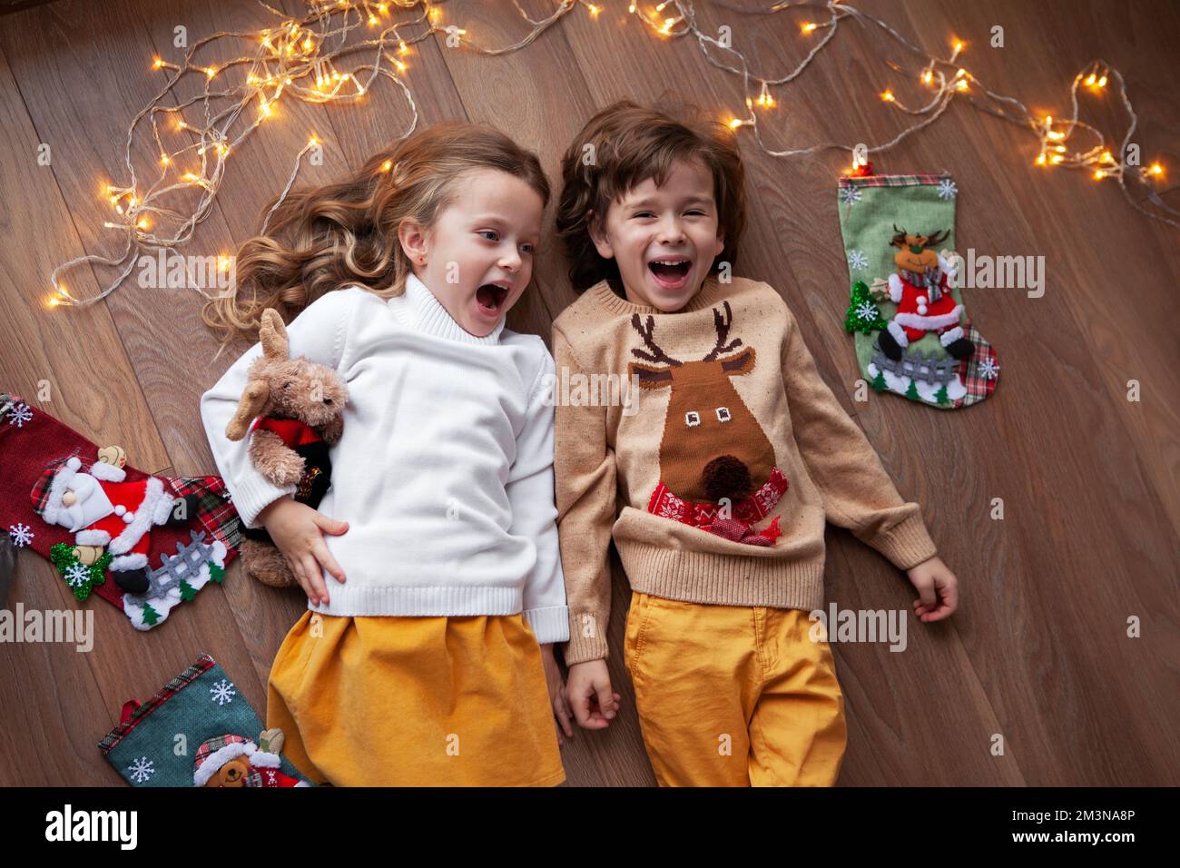 Kinder liegen zusammen, lachen und haben Spaß mit Weihnachtsdekor und Girlande aus Glühbirnen. Wunderschöne Kinder, die einen Pullover mit Hirsch tragen. Draufsicht, Stockfoto