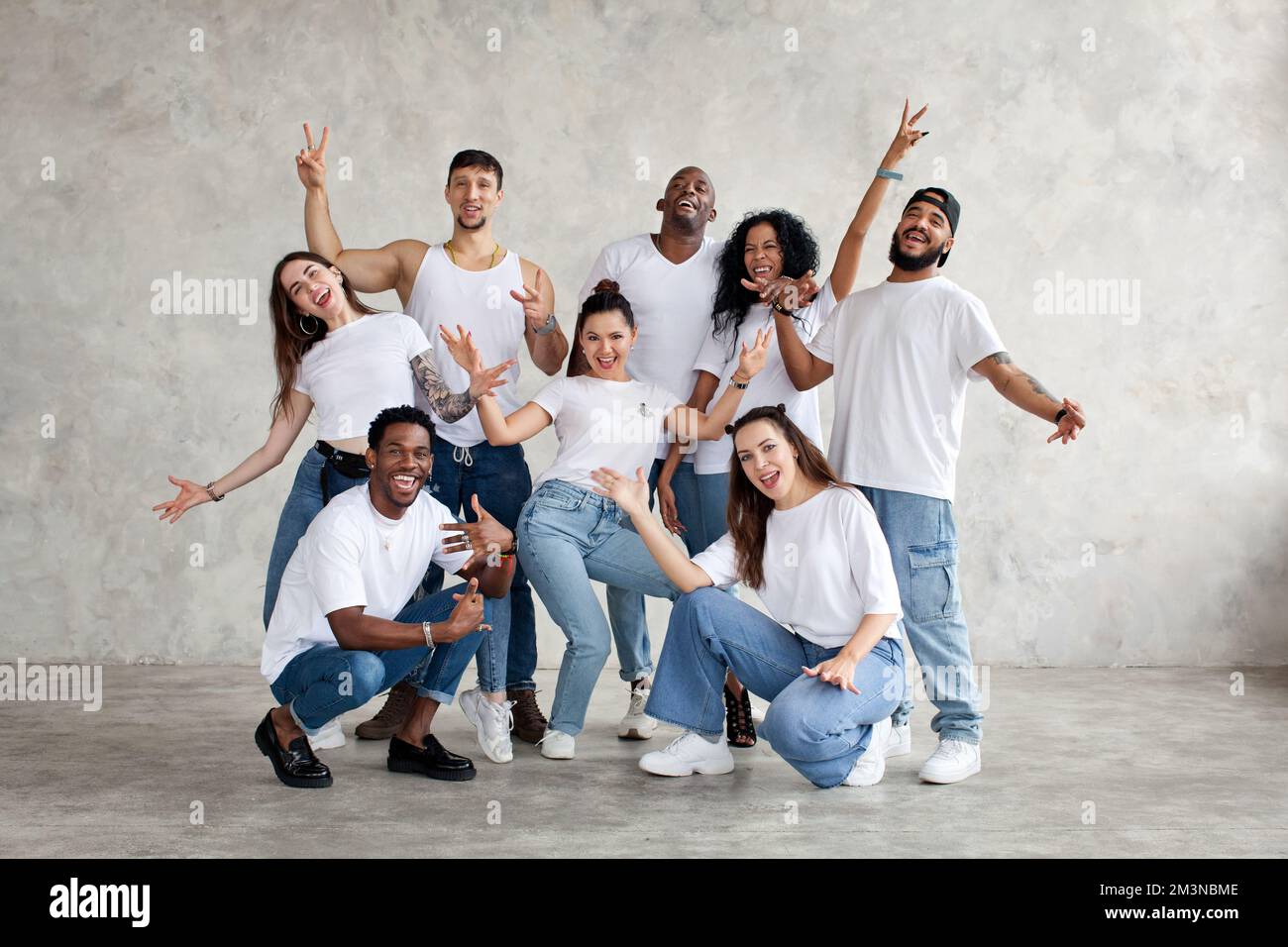 Multiethnische Gruppe glücklicher Freunde lächelt und hebt die Hände hoch. Verschiedene junge Menschen stehen zusammen in weißen Hemden und Jeans an der Hintergrundwand in St. Stockfoto