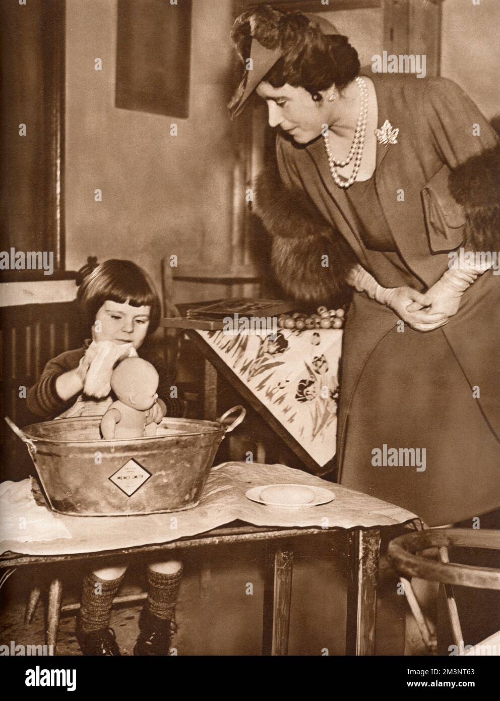 Die Königinmutter (1900-2002) mit evakuierten Kindern aus Battersea. Ein kleines vierjähriges Mädchen namens Shirely hier spielt "Mütter" und reagiert begeistert auf den Vorschlag der Königinmutter, die Puppe hinter den Ohren zu waschen. Datum: 1939 Stockfoto