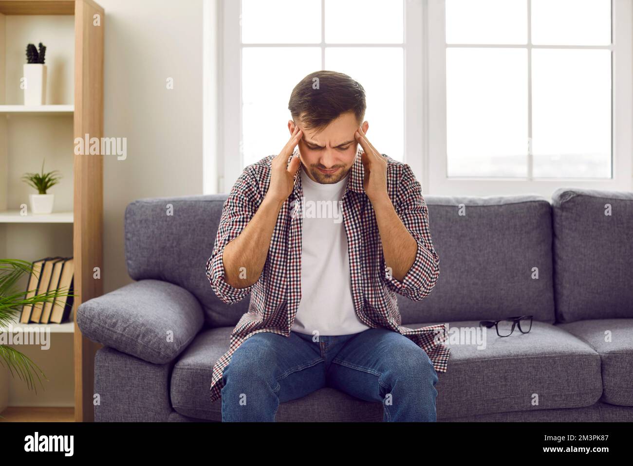 Der junge Mann, der sich die Schläfen reibt, leidet an Migräne, während er zu Hause auf der Couch sitzt. Stockfoto