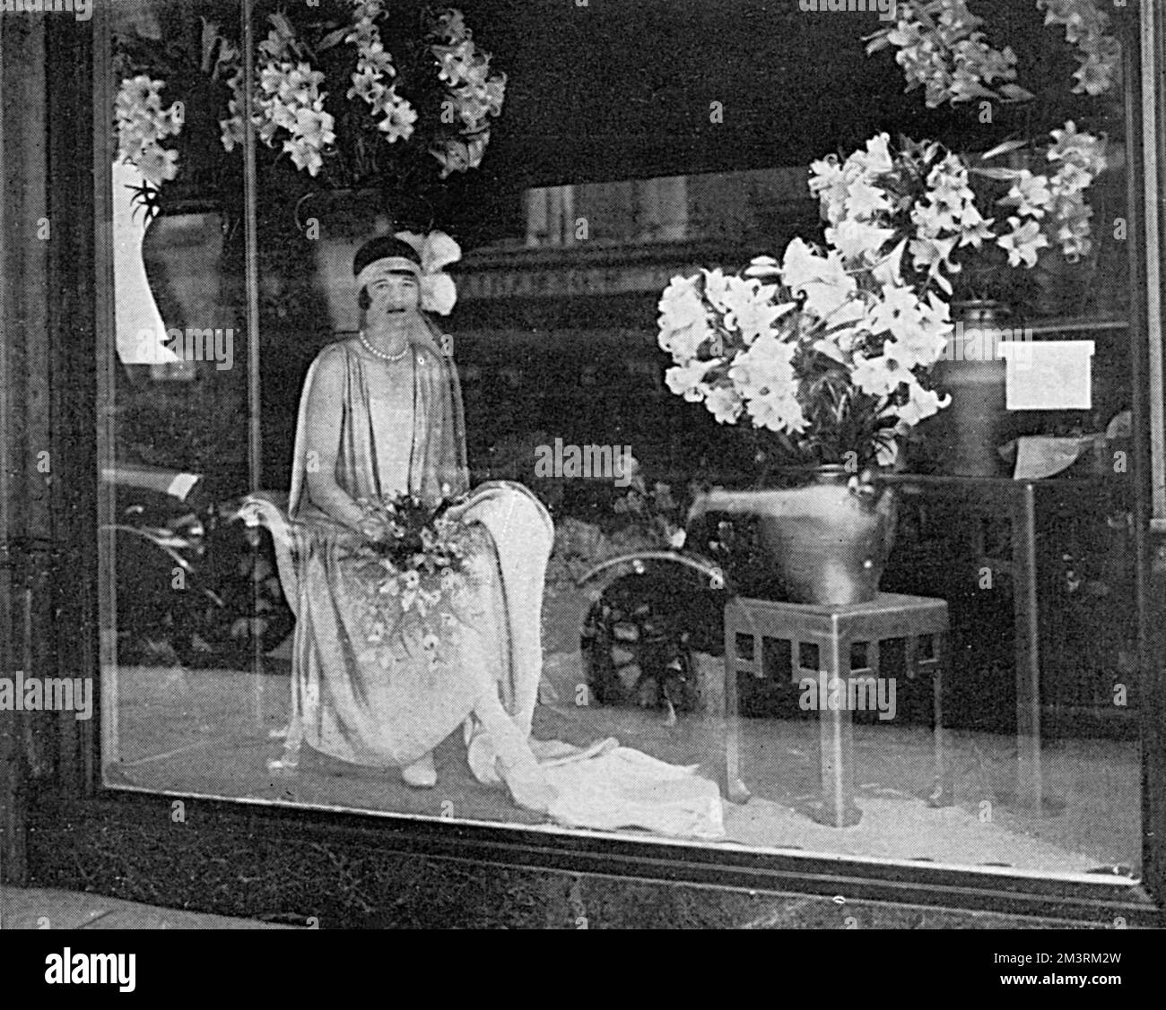 Eine neuartige Schaufensterausstellung in einem Geschäft in Knightsbridge, London, während der Sommersaison, zeigt eine junge Frau in einem Hofkleid mit Blumenstrauß und die traditionellen Federn, die für Hofpräsentationen getragen werden. Datum: 1928 Stockfoto