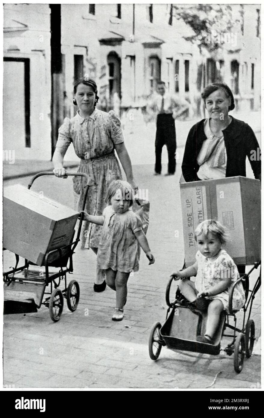 Zwei East End Mütter, die ihre Kinder Rollen und neue Baby-Gasmasken durch eine Londoner Straße. Begleitet von einer Beschriftung, die den Leser daran erinnert, immer eine Gasmaske bei sich zu tragen, und die Werbung für die Verfügbarkeit von Atemschutzgeräten für Babys macht. 1939 Stockfoto