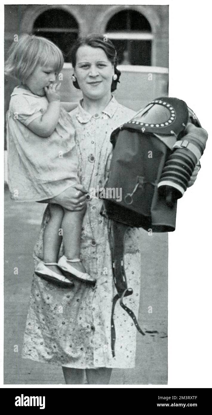 Eine Mutter im East End lächelt, während sie ihr Baby und ihre Baby-Gasmaske in jeder Hand hält. In der Zwischenzeit kommentiert die Kugel die "unverbindliche" Skepsis, mit der das Kind auf das Gerät starrt. September 1939 Stockfoto